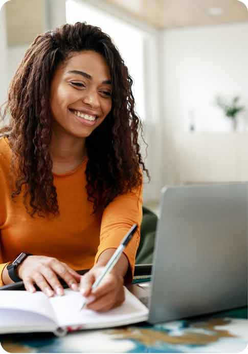 Young woman writing in notebook while looking at laptop
